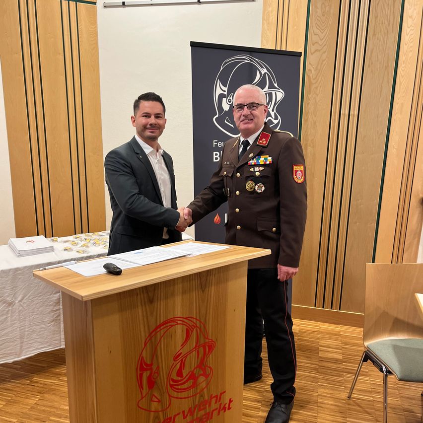 Zwei Männer schütteln sich die Hände vor einem Podium. Der Mann in Uniform trägt Medaillen und hält einen Stift. Ein Banner ist hinter ihnen.