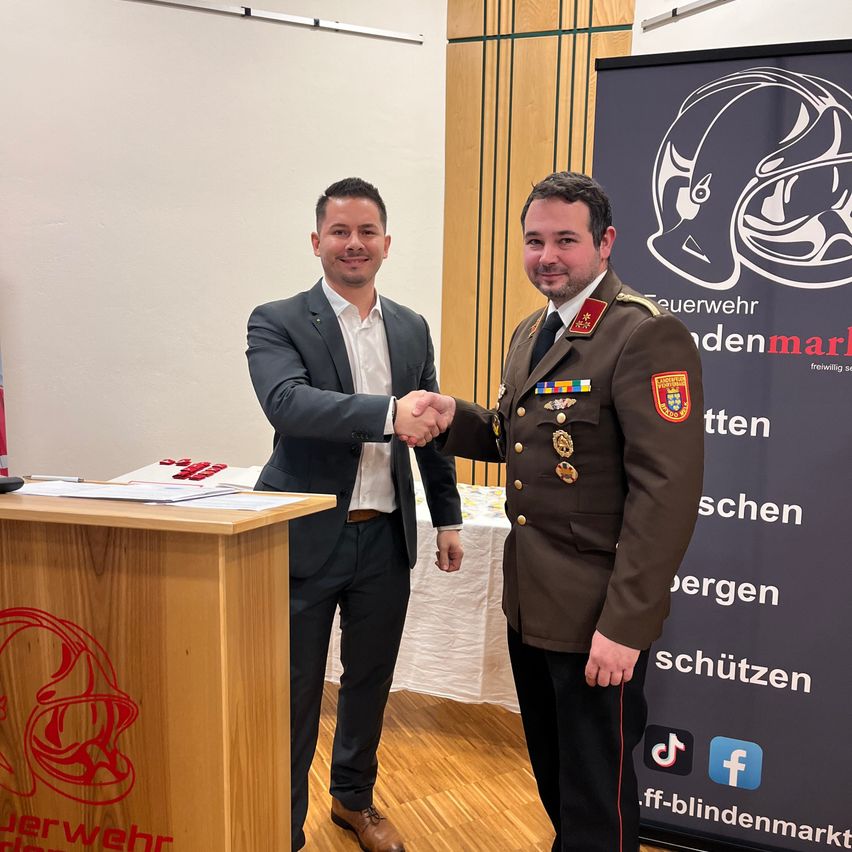 Zwei Männer in formeller Kleidung stehen lächelnd vor einem Podium mit einem roten Logo und schütteln sich die Hände. Dahinter befindet sich ein großes Banner mit einem militärischen Abzeichen und Text.