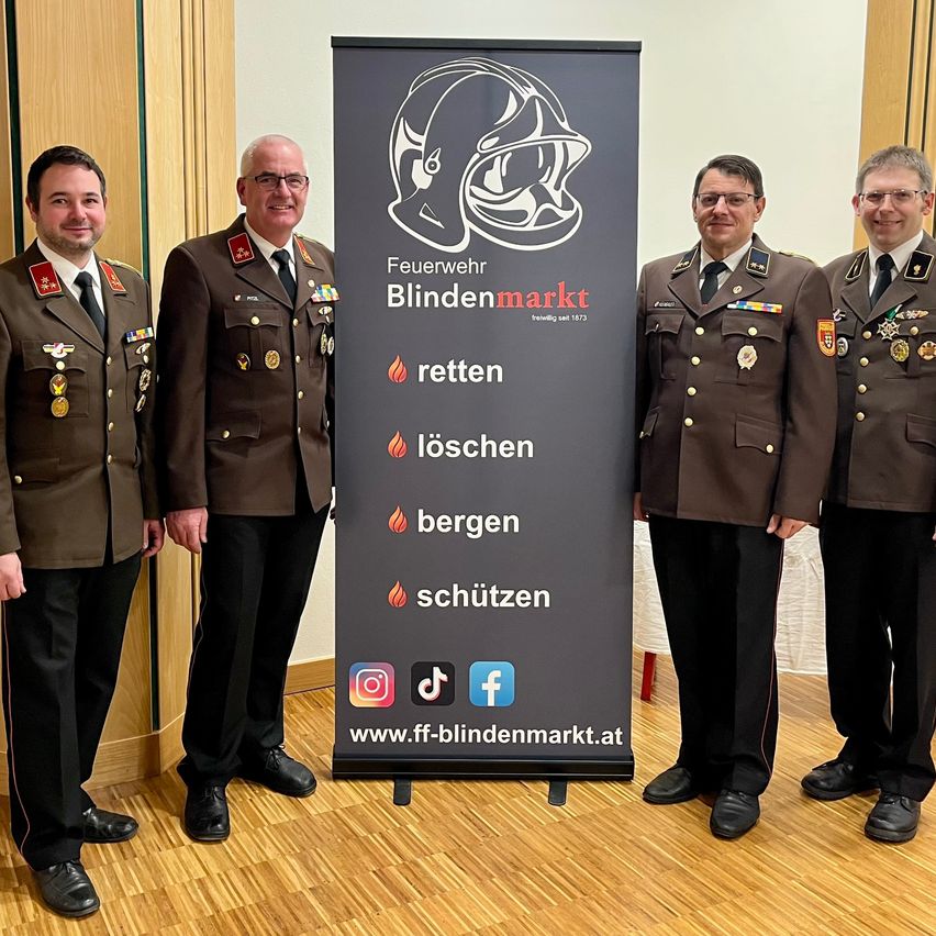 Vier Männer in Militäruniformen stehen neben einem Schild mit einem Helm-Logo, roten Flammen und Text auf Deutsch. Sie posieren für ein Foto in einem Raum mit Holzboden.