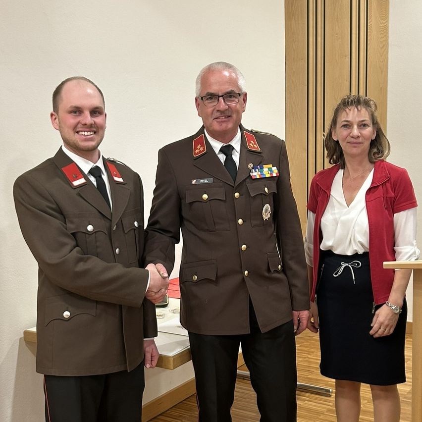Drei Menschen in Uniformen stehen nah beieinander und lächeln. Ein Mann gibt einem Militäroffizier die Hand. Eine Frau in einem roten Cardigan steht neben ihnen. Hinter ihnen ist eine Holzwand.