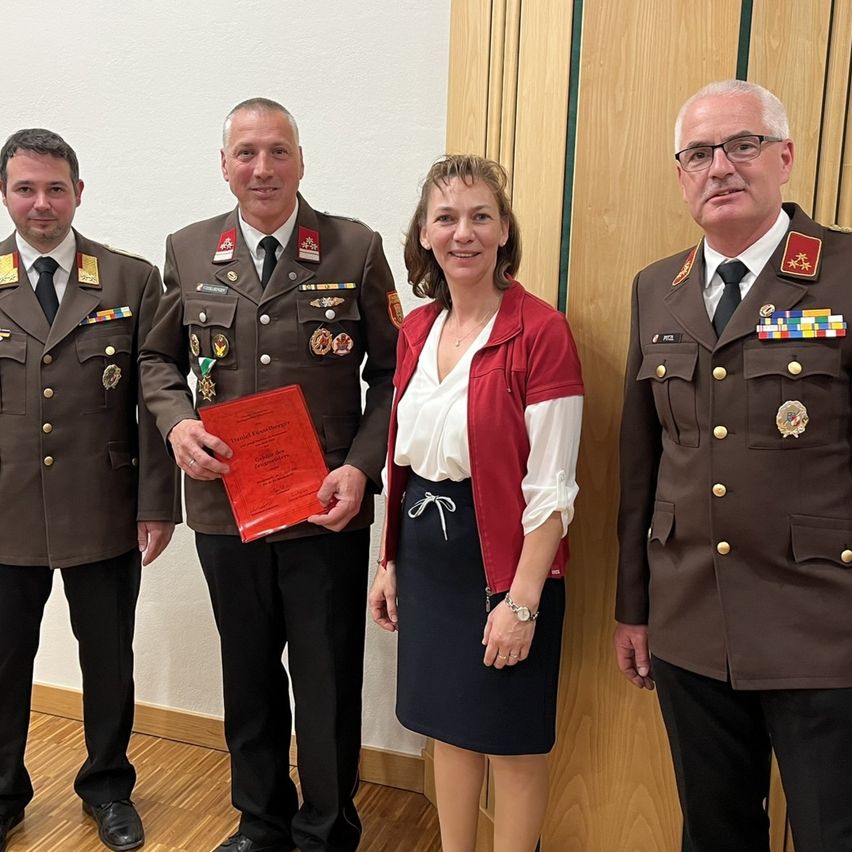 Vier Personen stehen zusammen. Eine Frau hält ein rotes Zertifikat. Die Männer tragen Militäruniformen und Medaillen. Sie stehen vor einer Holzwand.