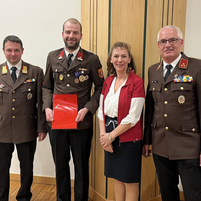Vier Menschen, zwei Männer und zwei Frauen, stehen zusammen. Die Männer sind in Militäruniformen und einer hält ein rotes Zertifikat. Die Frau in einer roten Weste steht in der Mitte und lächelt. Der Mann auf der rechten Seite trägt eine Brille.