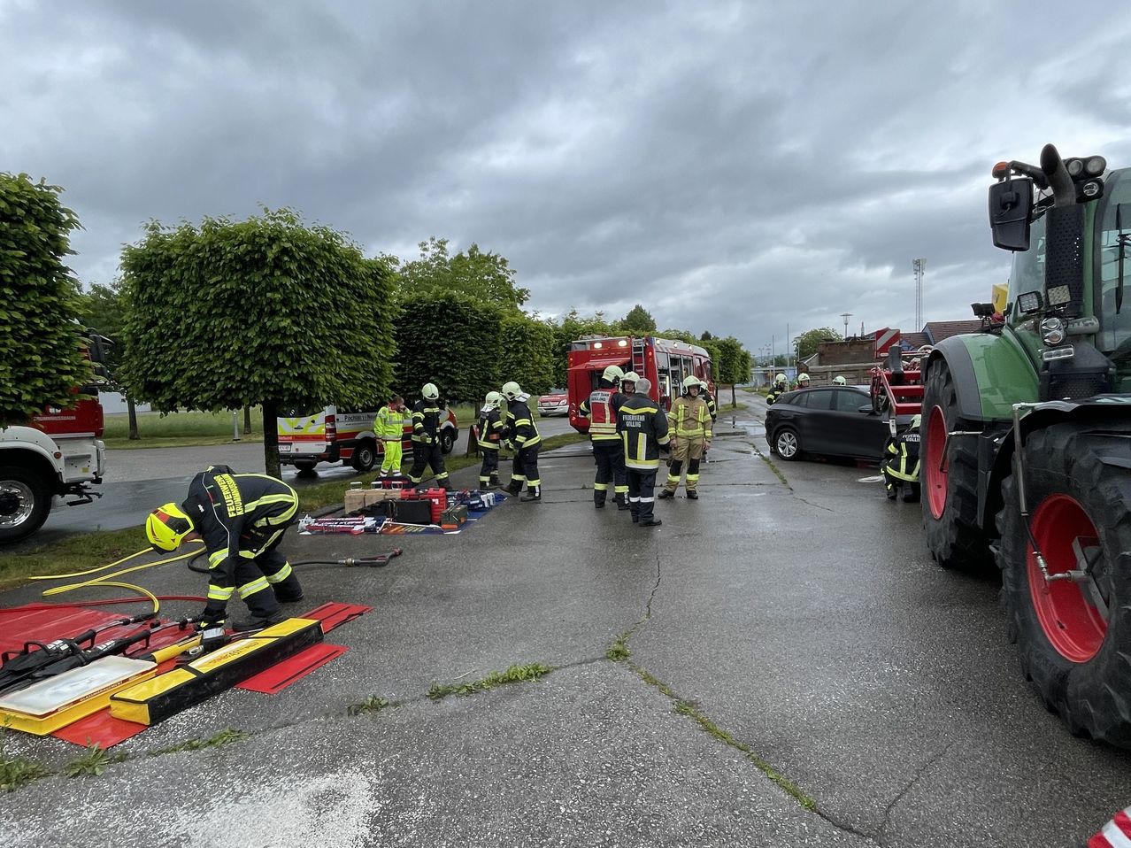 Firefighters in yellow and black uniforms work on equipment on a wet road, with fire trucks and a black car nearby.