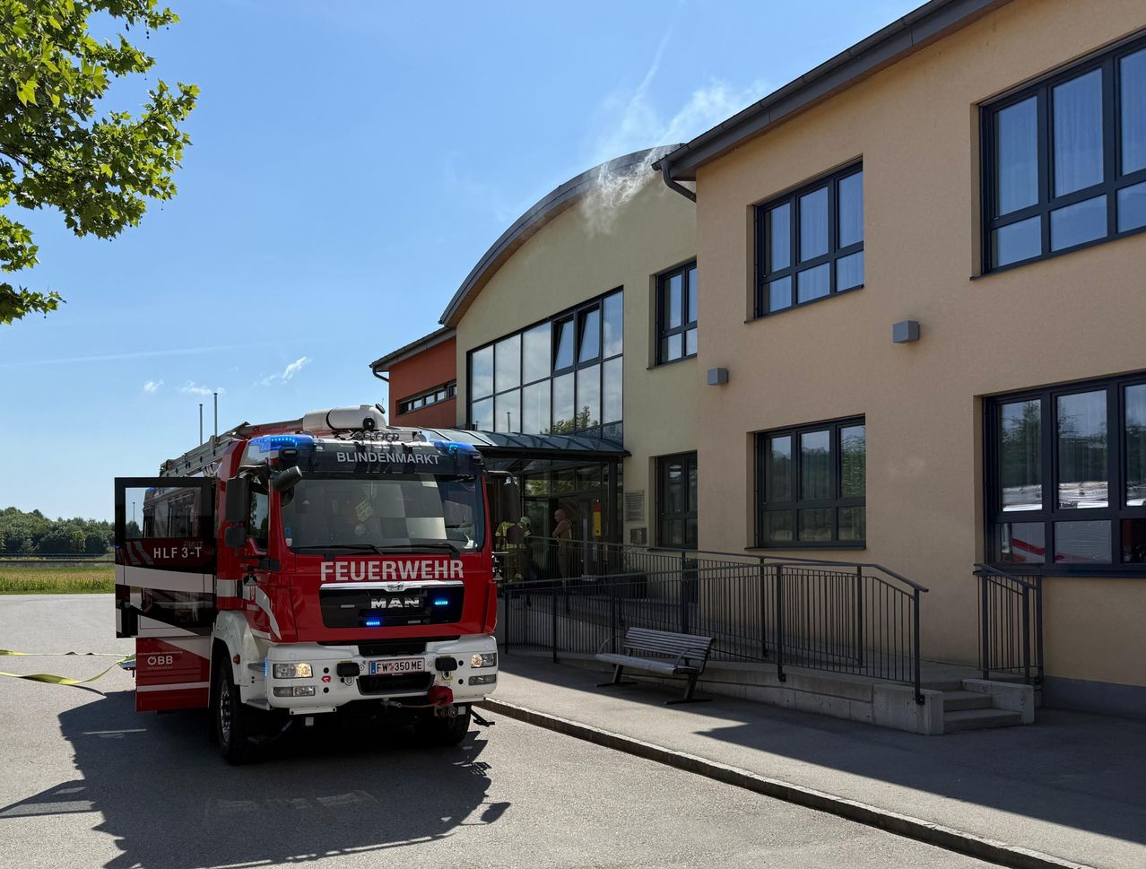 Ein roter Feuerwehrwagen mit dem Wort 'Feuerwehr' steht vor einem Gebäude mit Glasfenstern und einer Bank an der Seite.