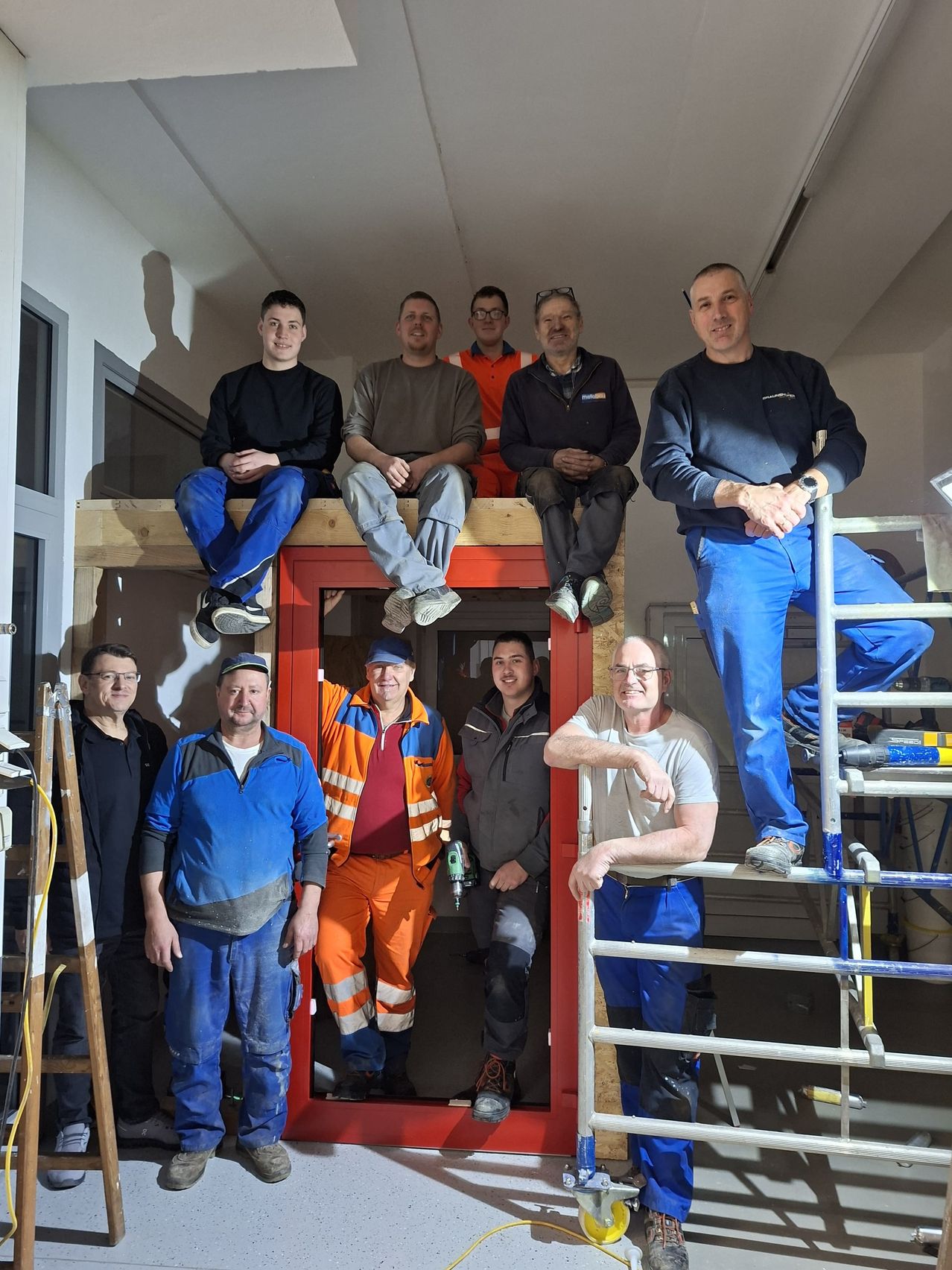 Eine Gruppe von Männern in Arbeitskleidung steht vor einer roten Tür. Sie lächeln und posieren für ein Foto. Hinter ihnen befindet sich eine weiße Wand, ein Fenster und eine Metallleiter. Sie sind wahrscheinlich Arbeiter auf einer Baustelle.