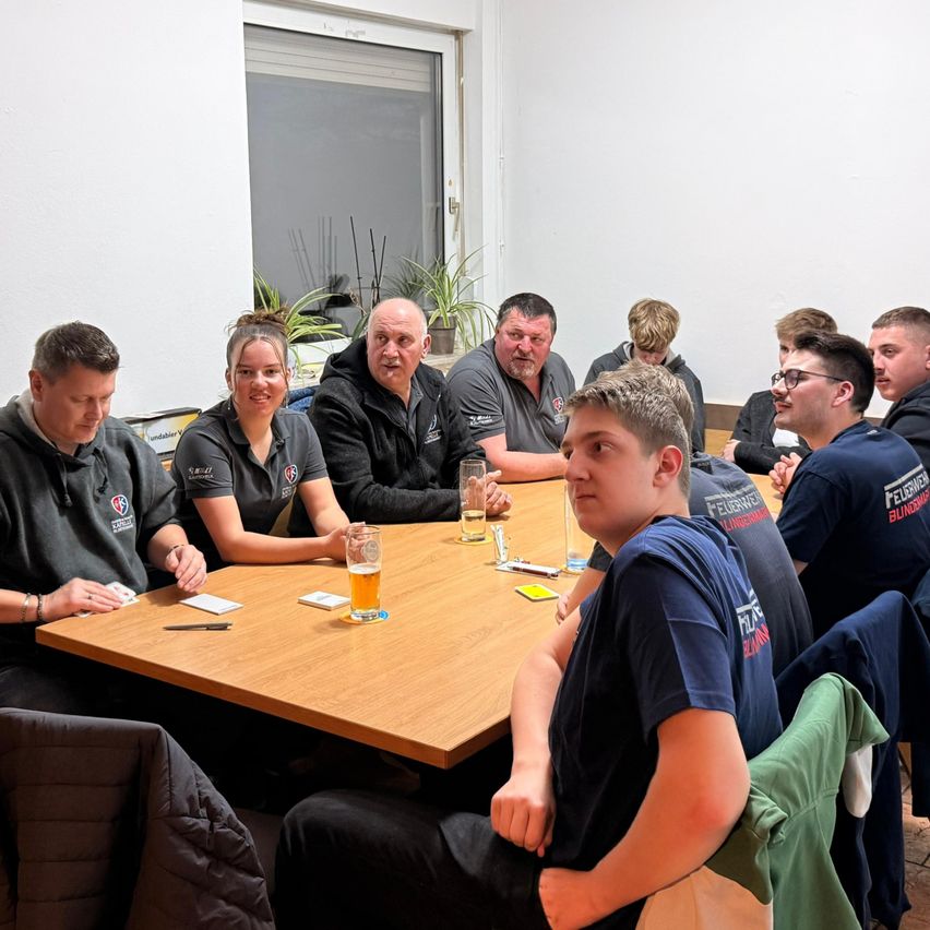 Eine Gruppe von Menschen in blauen und schwarzen Jacken sitzt um einen Tisch in einem Raum mit einer weißen Wand und einem Fenster. Einige haben Brillen und andere haben Biergläser.
