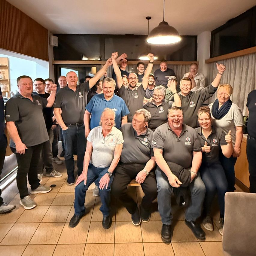 Eine Gruppe von Menschen in Uniform, einschließlich Männer und Frauen, posiert für ein Foto mit einigen erhobenen Händen in einem Raum mit gefliestem Boden und einer Lampe.