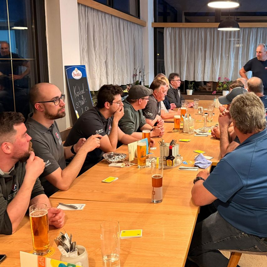 Mehrere Männer sitzen um einen Tisch mit Gläsern Bier. Ein Mann steht neben einer Tafel. Sie scheinen eine Besprechung zu führen.