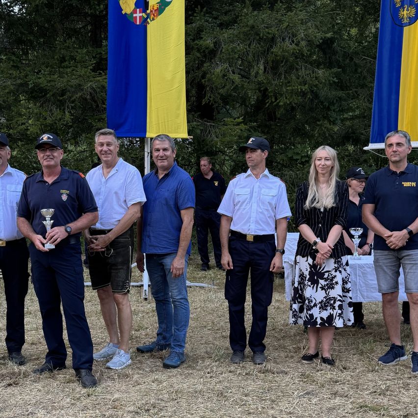 Eine Gruppe von Menschen steht auf einem Feld mit zwei Fahnen im Hintergrund. Sie alle lächeln und halten Pokale.