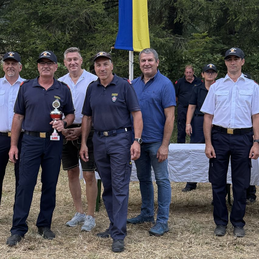 Eine Gruppe von Männern in Uniform steht auf einem Außengelände, einer hält eine Trophäe, mit einer Flagge und einem Tisch im Hintergrund.