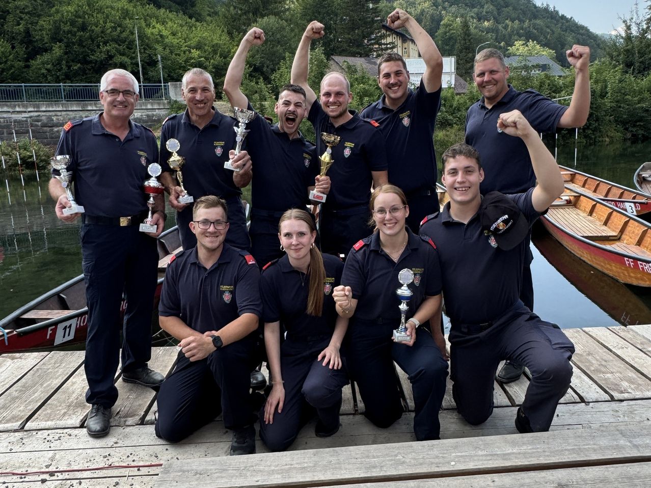Eine Gruppe von Feuerwehrleuten in Uniform, die lächelnd und mit ihren Trophäen auf einem Holzsteg in der Nähe eines Gewässers für ein Foto posieren.