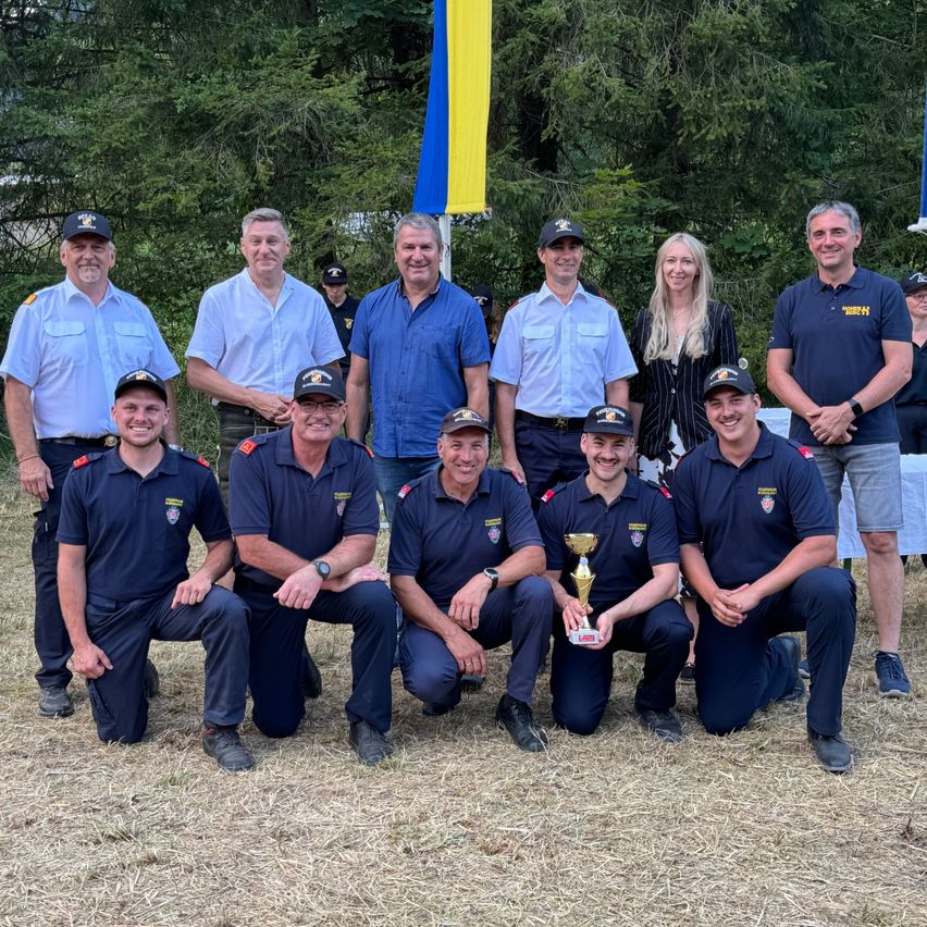 Eine Gruppe von Feuerwehrleuten und Offiziellen posiert mit einem Pokal auf einem Feld. Im Hintergrund befinden sich Bäume und eine Flagge.