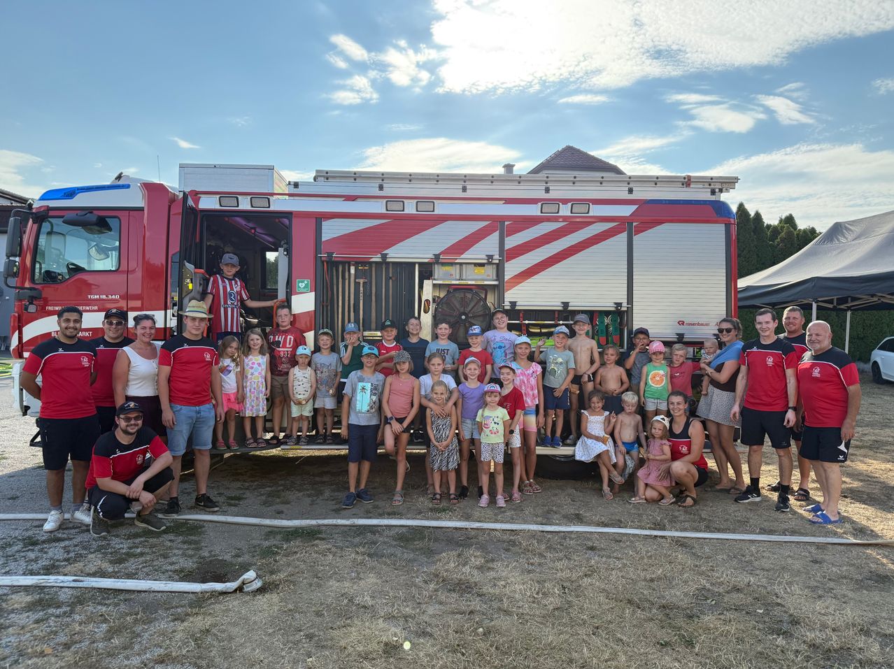 Eine Gruppe von Kindern und Erwachsenen posiert für ein Foto vor einem Feuerwehrauto an einem sonnigen Tag.