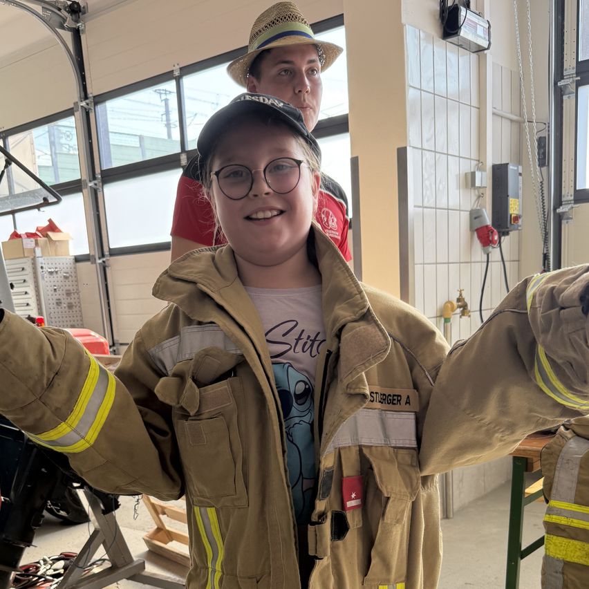 Ein junger Mensch in einer Feuerwehruniform posiert mit ausgestreckten Armen in einer Garage, mit einer Person in einem Hut hinter ihm.