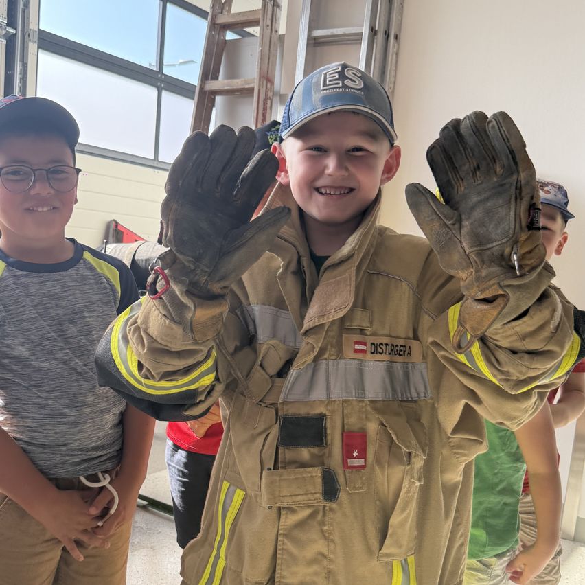 Ein junger Junge in einer Feuerwehruniform, lächelt und zeigt seine Handschuhe, mit einem anderen Jungen mit Brille und einem dritten Kind im Hintergrund.