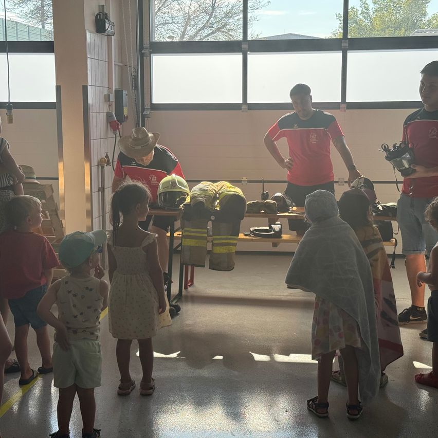 Eine Gruppe von Kindern und Erwachsenen versammelt sich um einen Tisch mit Feuerwehrausrüstung. Zwei Männer in roten und schwarzen Uniformen stehen hinter dem Tisch.