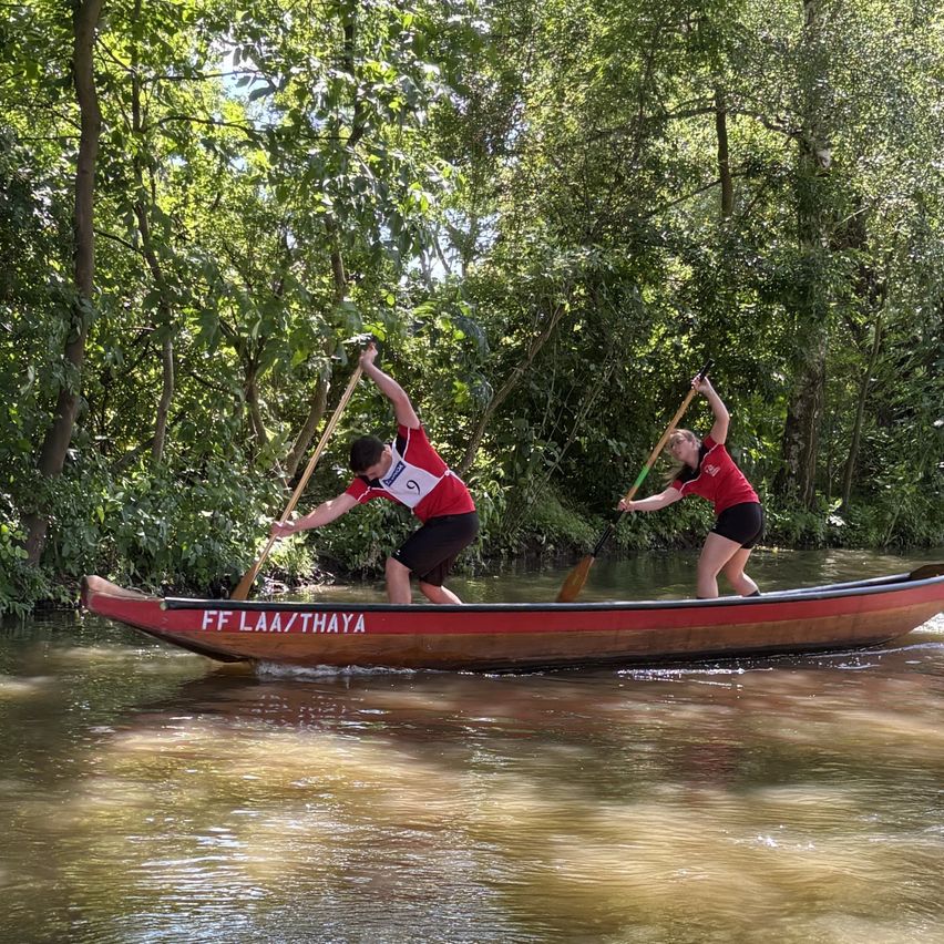 Zwei Personen paddeln ein Kanu mit der Aufschrift FF LAA/THAYA auf einem Fluss, umgeben von Bäumen. Beide tragen rote Shirts, Shorts und eine Nummer 5 auf ihren Shirts.