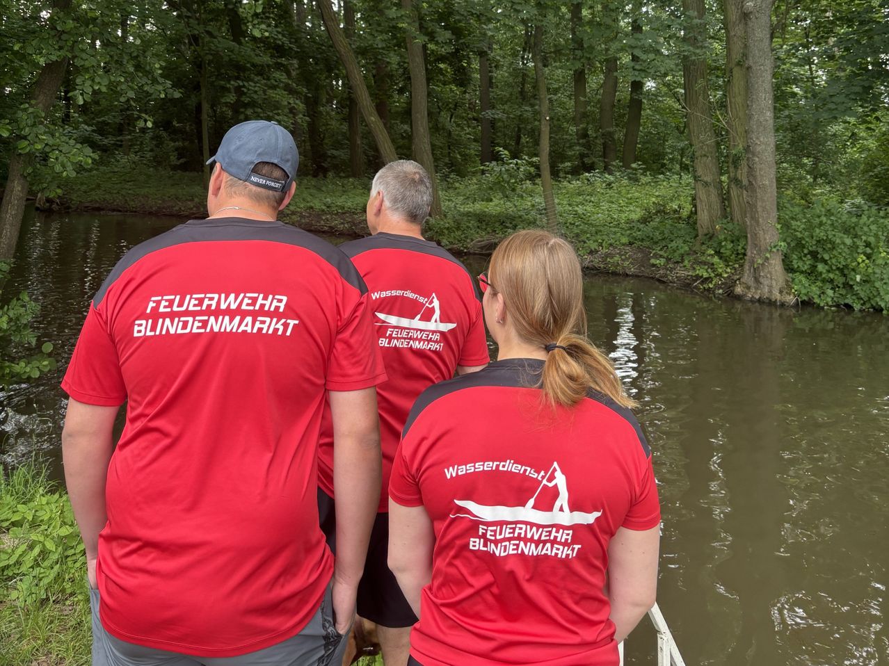 Drei Menschen stehen am Fluss. Alle tragen rote T-Shirts mit weißem Text. Zwei Männer stehen nebeneinander, und eine Frau steht vor ihnen. Bäume säumen das Flussufer.