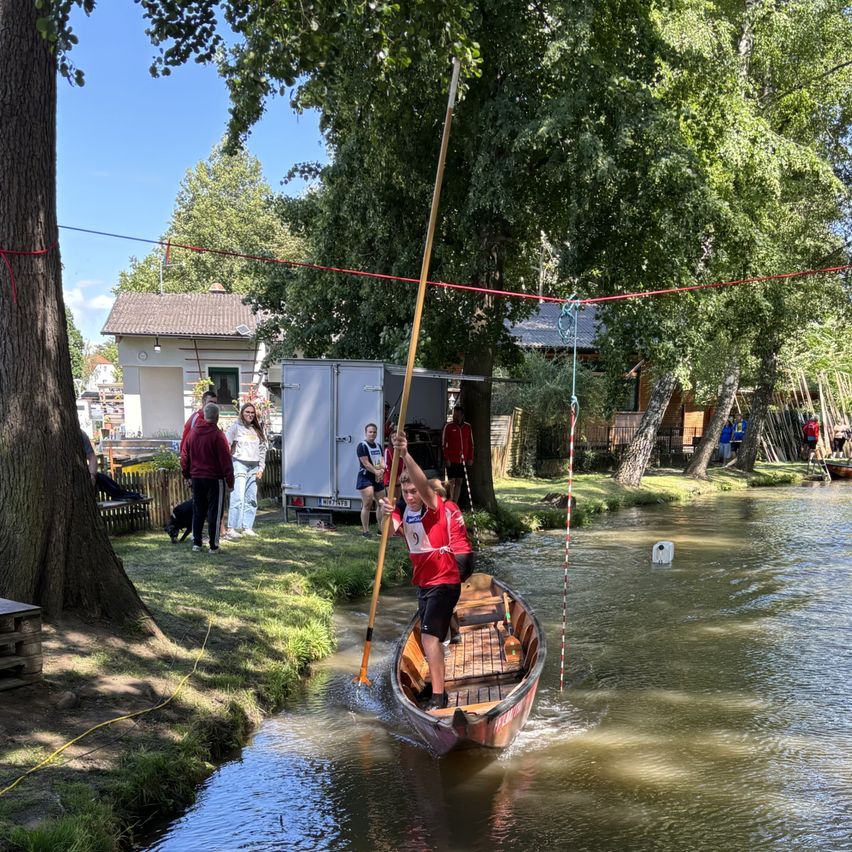 Ein junger Mann in einem roten Shirt steht in einem kleinen Boot auf einem Fluss und hält eine Ruder. Zuschauer beobachten vom Grasufer aus. Bäume und Häuser sind im Hintergrund.
