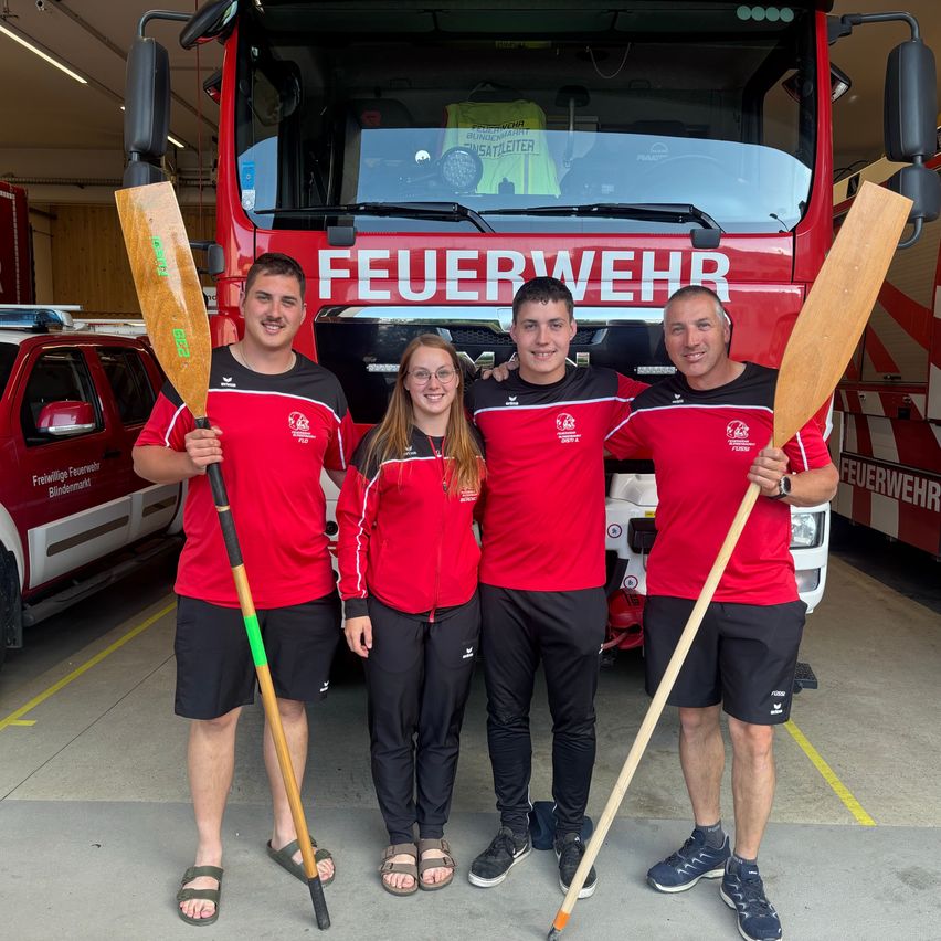 Vier Personen in roten und schwarzen Uniformen posieren für ein Foto vor einem Feuerwehrwagen. Drei von ihnen halten Holzpaddel in der Hand. Der Feuerwehrwagen steht in einer Garage.