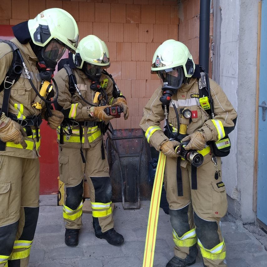 Drei Feuerwehrleute in Schutzausrüstung stehen außerhalb. Sie halten Geräte und scheinen zu diskutieren. Dahinter befindet sich eine Ziegelmauer.