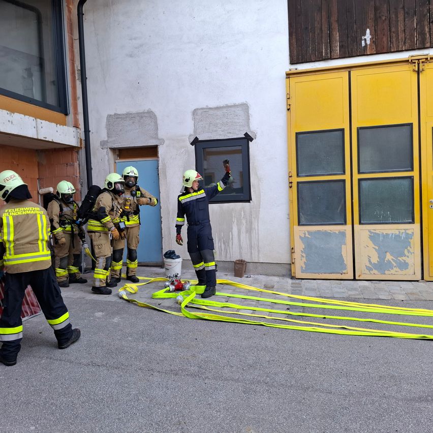 Feuerwehrleute in gelben Uniformen bereiten sich auf einen Notfall vor. Einer zeigt auf ein Fenster, während andere neben einem gelben Schlauch auf der Straße stehen.