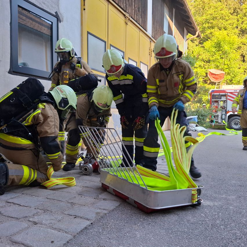 Eine Gruppe Feuerwehrleute, ausgestattet mit Helmen und Sicherheitsanzügen, trainiert vor einem Gebäude und benutzt Werkzeuge an einem gelben Objekt.