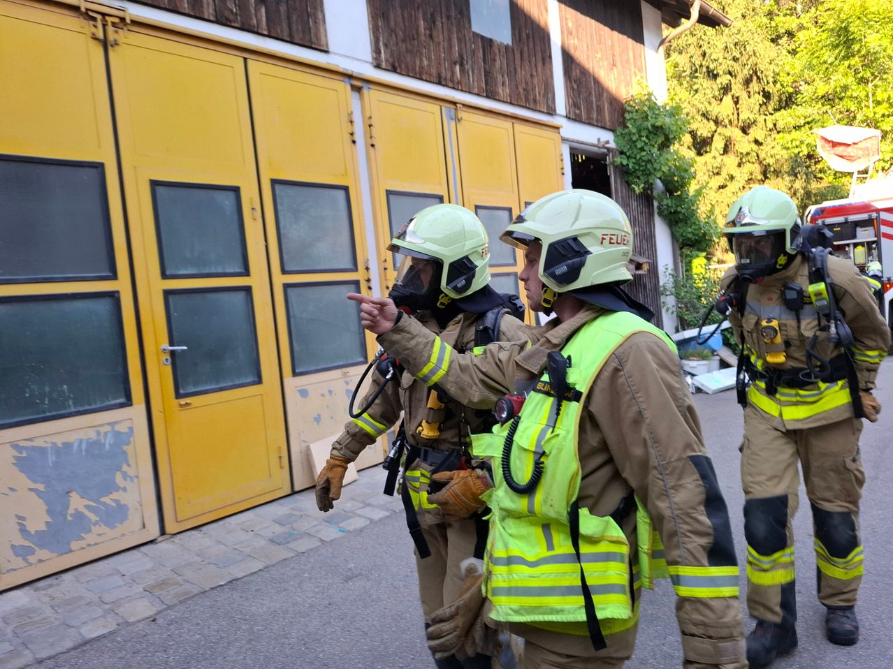 Drei Feuerwehrleute besprechen etwas, einer von ihnen zeigt auf das Gebäude mit gelben Türen.