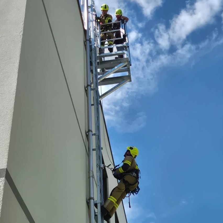 Drei Feuerwehrleute in Schutzkleidung klettern mit einer Metallleiter die Außenwand eines Gebäudes hinauf. Einer steht auf einer Plattform, während die anderen beiden aufsteigen.