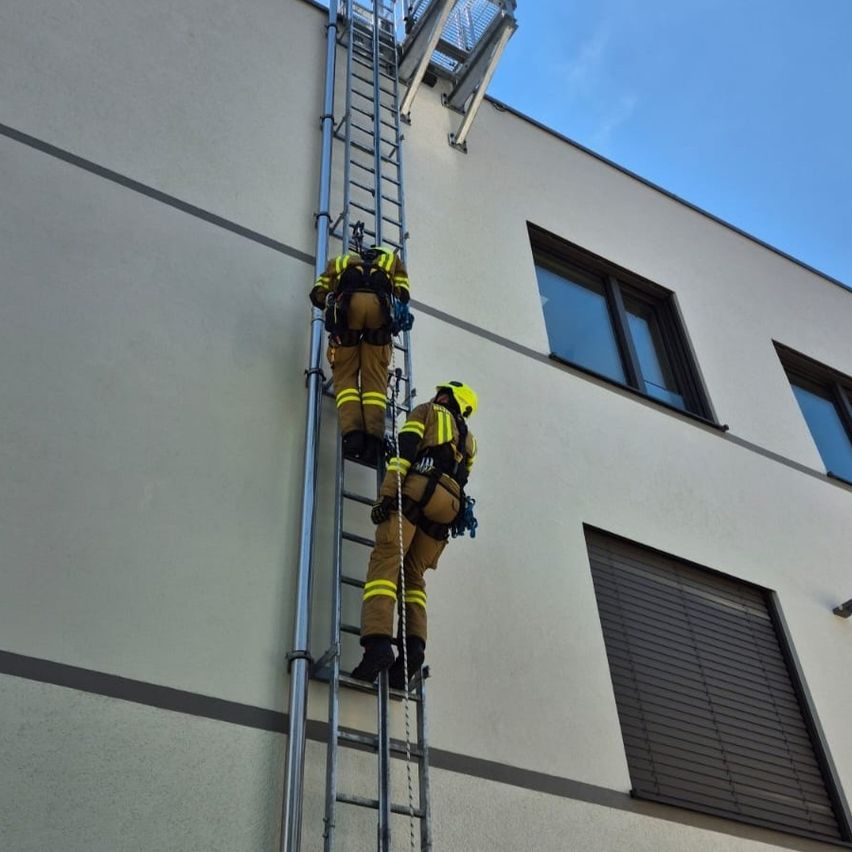 Zwei Feuerwehrleute in gelben Uniformen und Helmen klettern eine Leiter an einem Gebäude hoch. Einer ist in der Nähe der Spitze, der andere ist halbwegs oben. Das Gebäude hat eine Metallleiter und eine Metallplattform.