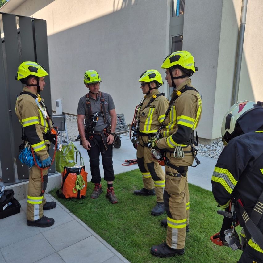 Feuerwehrleute in gelben und schwarzen Uniformen stehen auf einem Rasen, tragen Helme und unterhalten sich. Sie stehen vor einem Gebäude mit einem Trolley und einigen Ausrüstungsgegenständen.
