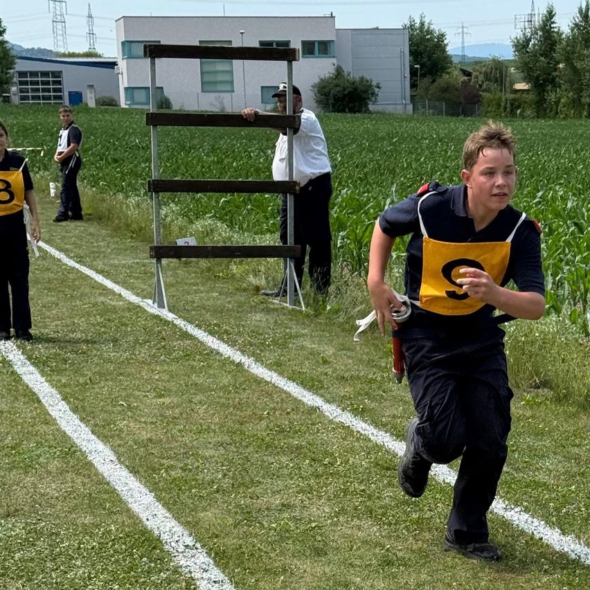 Ein junger Junge läuft in einem Rennen auf einem Grasfeld. Hinter ihm beobachten zwei Offizielle. Einer steht in der Nähe einer Leiter, während der andere einen Hut trägt.