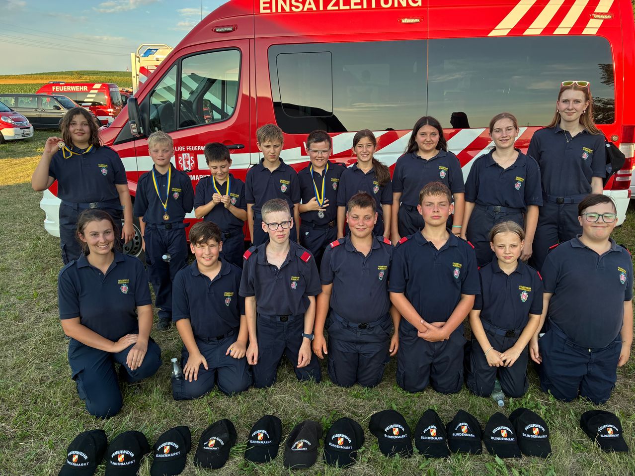 Eine Gruppe junger Feuerwehrleute in Uniform posiert vor einem Feuerwehrwagen. Sie alle lächeln, einige mit Medaillen um den Hals.