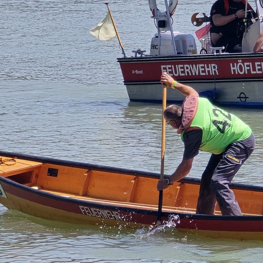 Ein Feuerwehrmann mit grüner Weste und der Nummer 27 steht in einem Boot und hält einen Stock. Ein anderer Feuerwehrmann sitzt in einem Boot mit der Aufschrift 'Feuerwehr Höfle'.