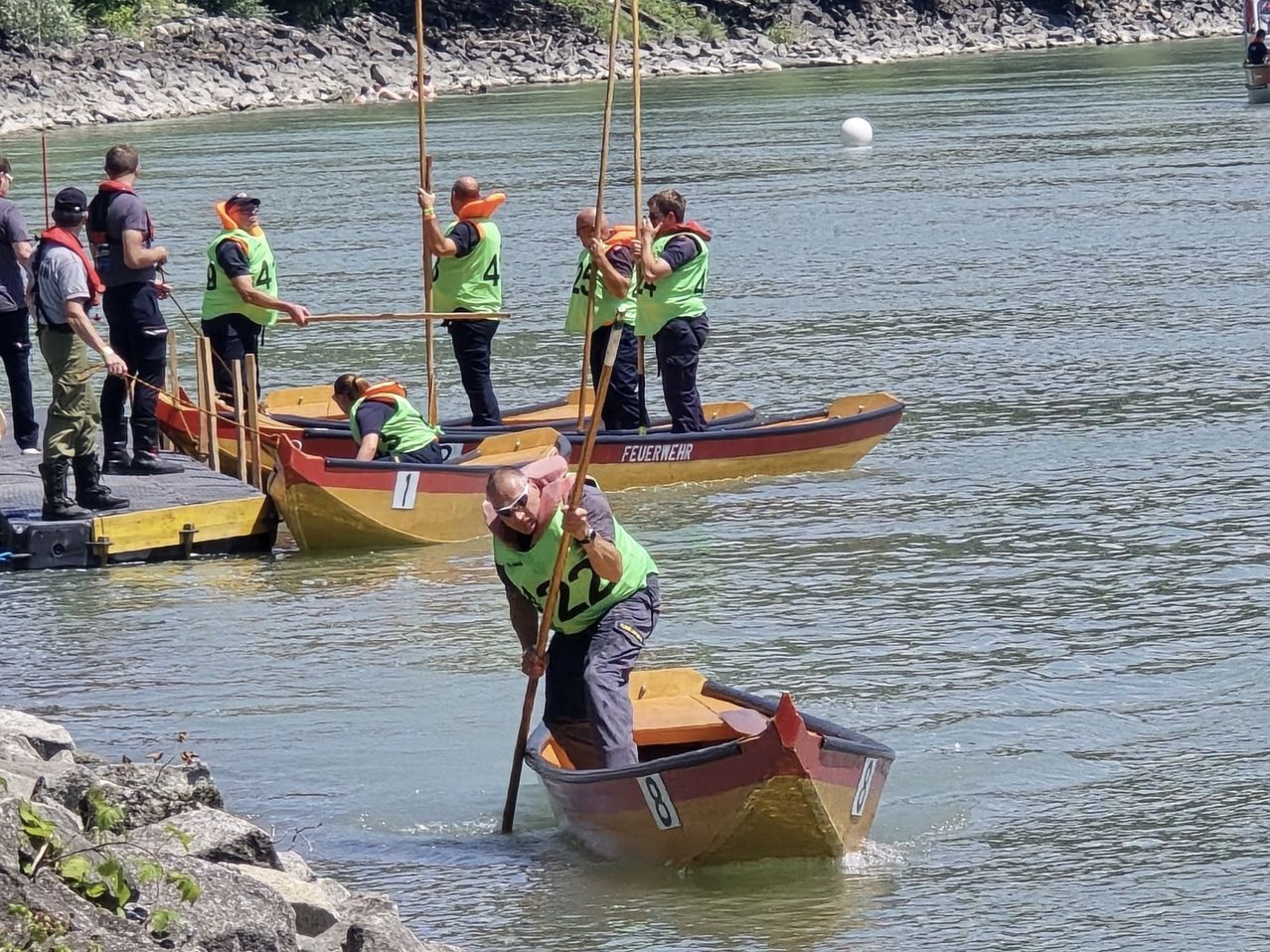 Fünf Personen in Schwimmwesten, die grüne Westen tragen, rudern auf Booten auf einem Fluss. Ein Mann im vorderen Boot trägt eine Sonnenbrille und paddelt.