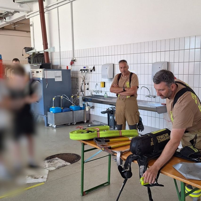 Zwei Feuerwehrleute bereiten Ausrüstung auf einem Tisch in einer Feuerwache vor. Einer hat eine Werkzeugtasche, während der andere in der Nähe steht. Eine Person im Hintergrund erscheint verschwommen.