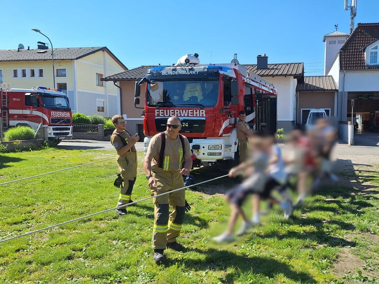 Ein Feuerwehrauto steht vor einem Haus. Feuerwehrleute stehen auf dem Gras mit Kindern. Die Kinder halten Seile, möglicherweise für eine Trainingsübung.