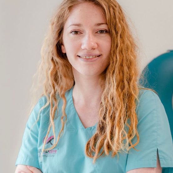 Eine Frau mit blonden Haaren lächelt und posiert für ein Foto in einem medizinischen Umfeld. Sie trägt ein blaues Hemd mit Logo.