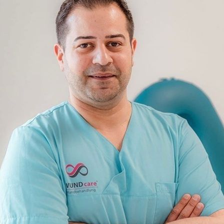 Ein lächelnder Mann in einem blauen Kittel steht mit verschränkten Armen. Er befindet sich in einem Krankenhaus. Hinter ihm ist ein blauer Ballon.