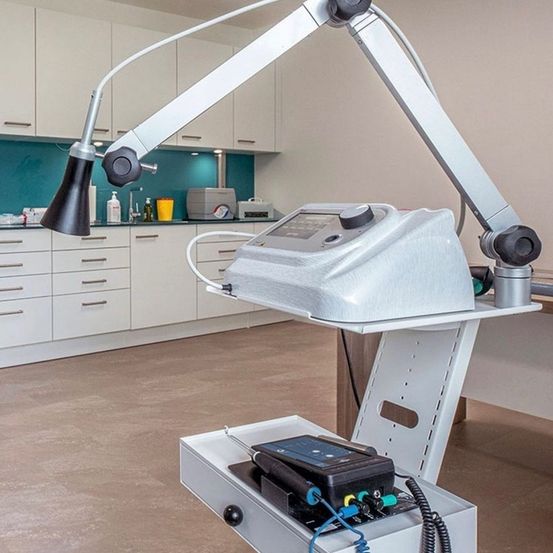 Ein Innenraum eines medizinischen oder zahnärztlichen Büros mit einem Metallarm, der ein Gerät hält. Ein Küchenbereich mit Schränken und einem Waschbecken ist im Hintergrund zu sehen.