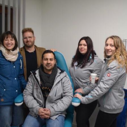 Fünf Menschen posieren für ein Foto. Der Mann in der Mitte sitzt auf einem blauen Stuhl. Die beiden Frauen rechts und links lächeln. Die Frau links hält eine Tasse. Die Frau in der Mitte hält eine Tasse. Der Mann links steht hinter dem Mann im Stuhl.
