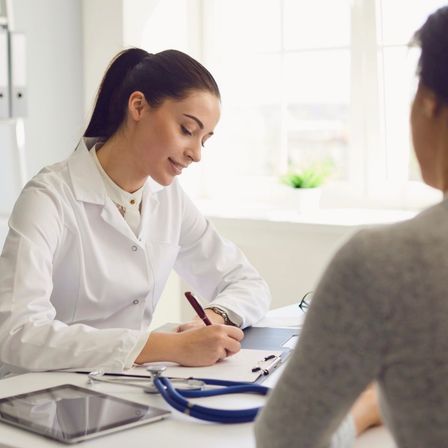 Eine Ärztin in einem weißen Laborkittel sitzt an einem Schreibtisch, lächelt und schreibt auf Papier. Ein Patient steht in der Nähe und hört aufmerksam zu. Auf dem Schreibtisch befinden sich ein Tablet, ein Stift und ein Stethoskop.