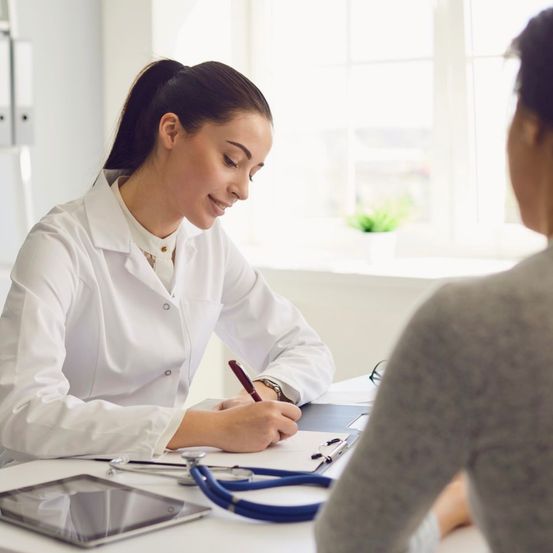 Eine Ärztin in einem weißen Laborkittel sitzt an einem Schreibtisch, lächelt und schreibt auf Papier. Ein Patient steht in der Nähe und hört aufmerksam zu. Auf dem Schreibtisch befinden sich ein Tablet, ein Stift und ein Stethoskop.