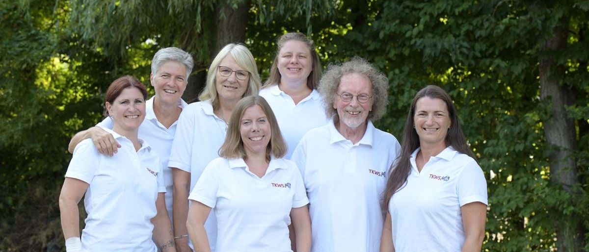 Eine Gruppe von Menschen in weißen Poloshirts mit Logos steht zusammen im Freien. Sie lächeln und posieren für ein Foto vor Bäumen.