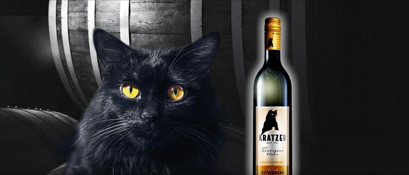 Eine schwarze Katze mit gelben Augen steht neben einer Flasche Weißwein namens Kratzer. Die Katze hat ein glänzendes schwarzes Fell und starrt nach vorne.