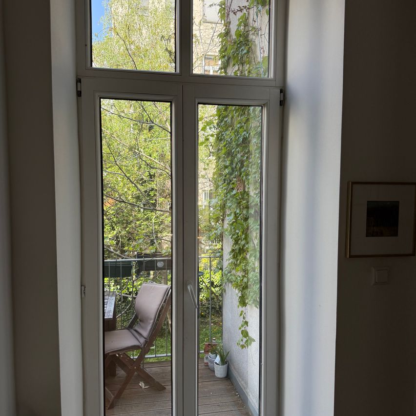 Eine offene Balkontür zeigt einen kleinen Holzbalkon mit einem gepolsterten Stuhl, einem Tisch und Blick auf einen üppigen grünen Baum. Der Balkon ist von einem Metallzaun umgeben und mit Kletterpflanzen geschmückt.