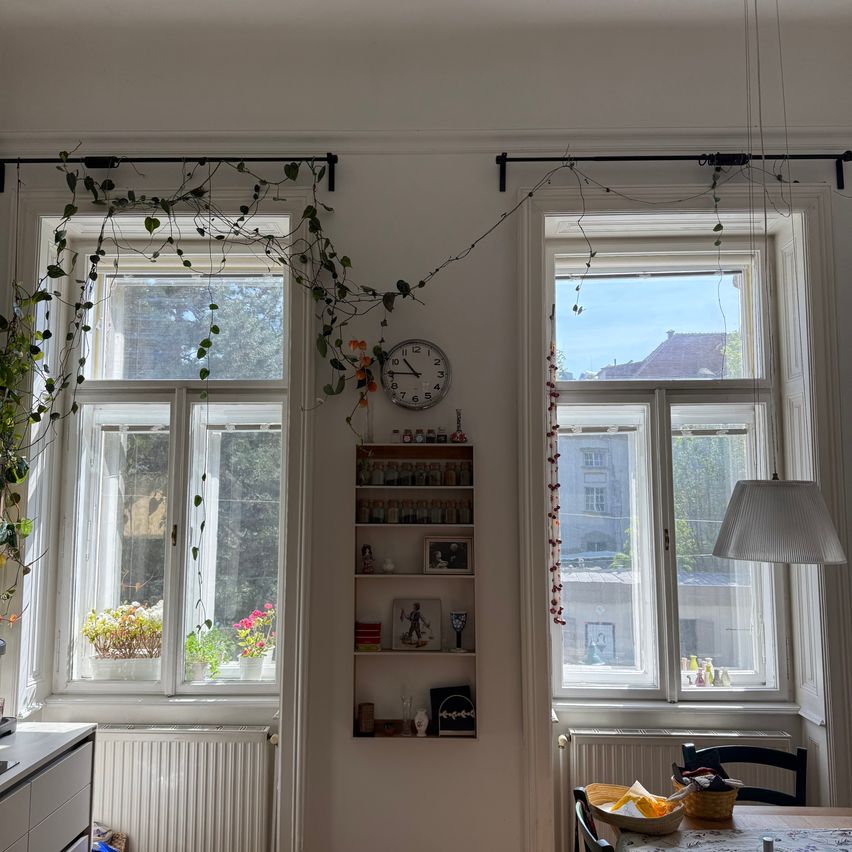 Ein Essbereich mit zwei großen Fenstern, eines mit hängenden Pflanzen und das andere mit einer Uhr. Die Wände sind weiß, und es gibt einen Tisch mit einer Schüssel und einer Lampe.