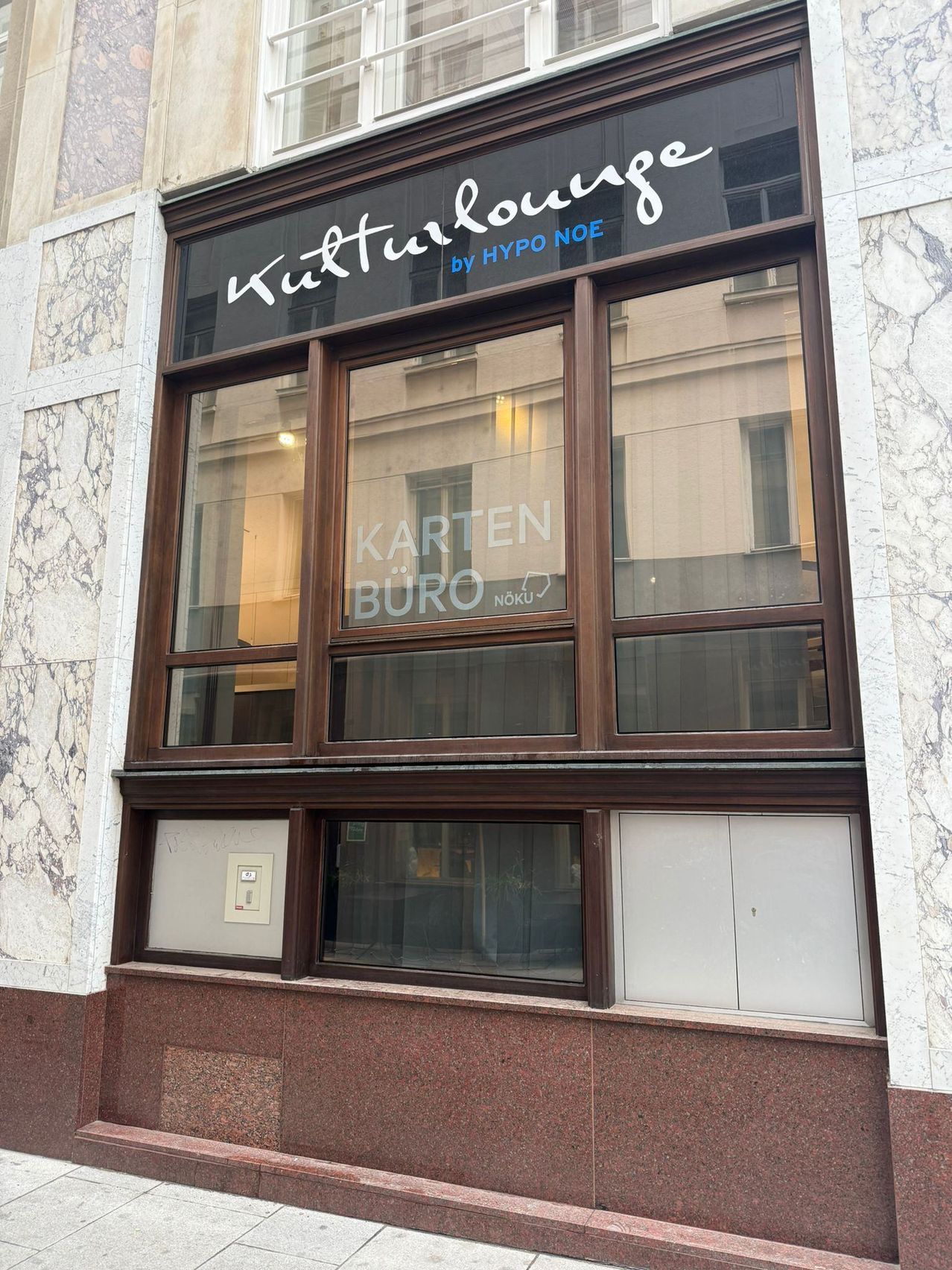 Die Schaufensterfront hat ein großes Fenster mit braunen Rahmen. Das Schild zeigt 'Titurllounge by HYPO NOE'. Unten ist ein Gebäude reflektiert. Im unteren Teil des Fensters steht 'Karten Buro Noku'.