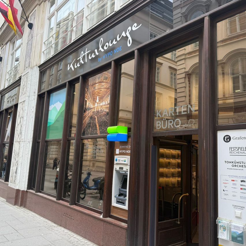 Ein Laden mit einem großen Schild, auf dem Tuturlounge by Hypno Noe steht. Es gibt eine Glastür mit einem Aufkleber für Karten Buro. Die Fenster des Ladens spiegeln ein nahes Gebäude, eine Flagge und einen Roller wider.