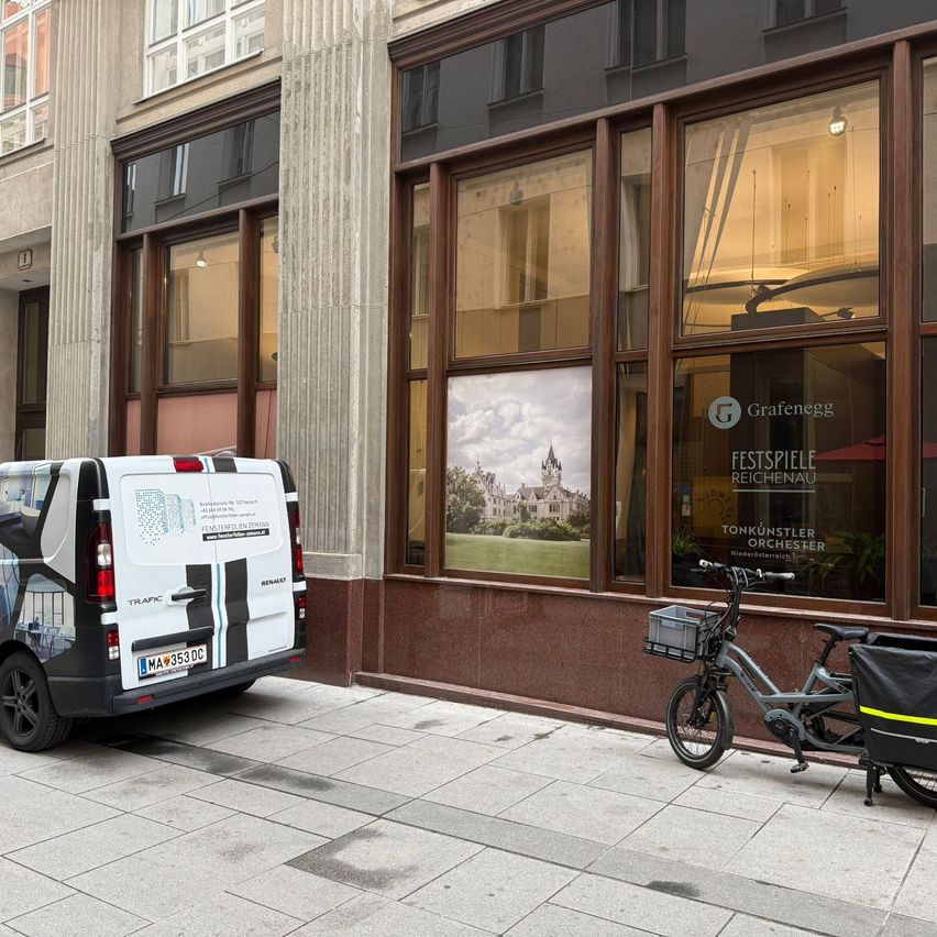 Ein weißer Lieferwagen steht auf dem Bürgersteig vor einem Gebäude. In der Nähe ist ein Fahrrad mit einem Korb davor geparkt. Das Gebäude hat große Glasfenster mit einem Bild einer Burg.